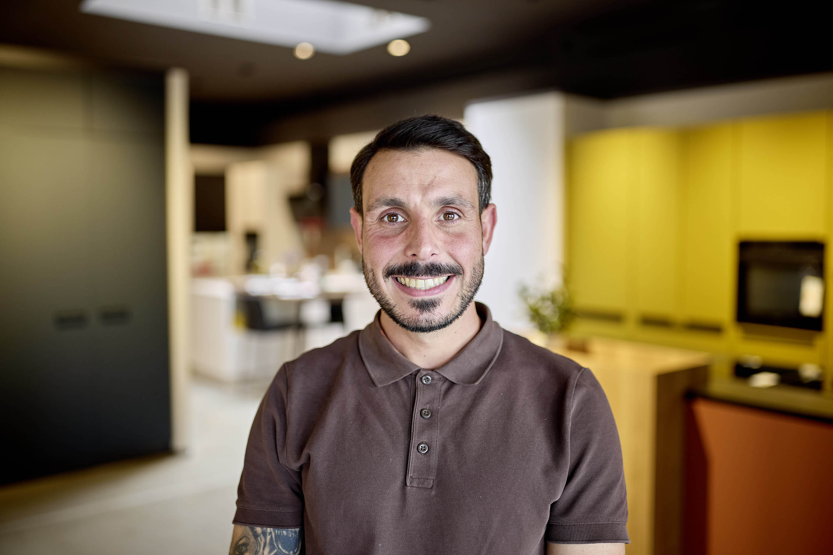 Eine lächelnde Person in einem braunen Poloshirt in einer modernen Küche.