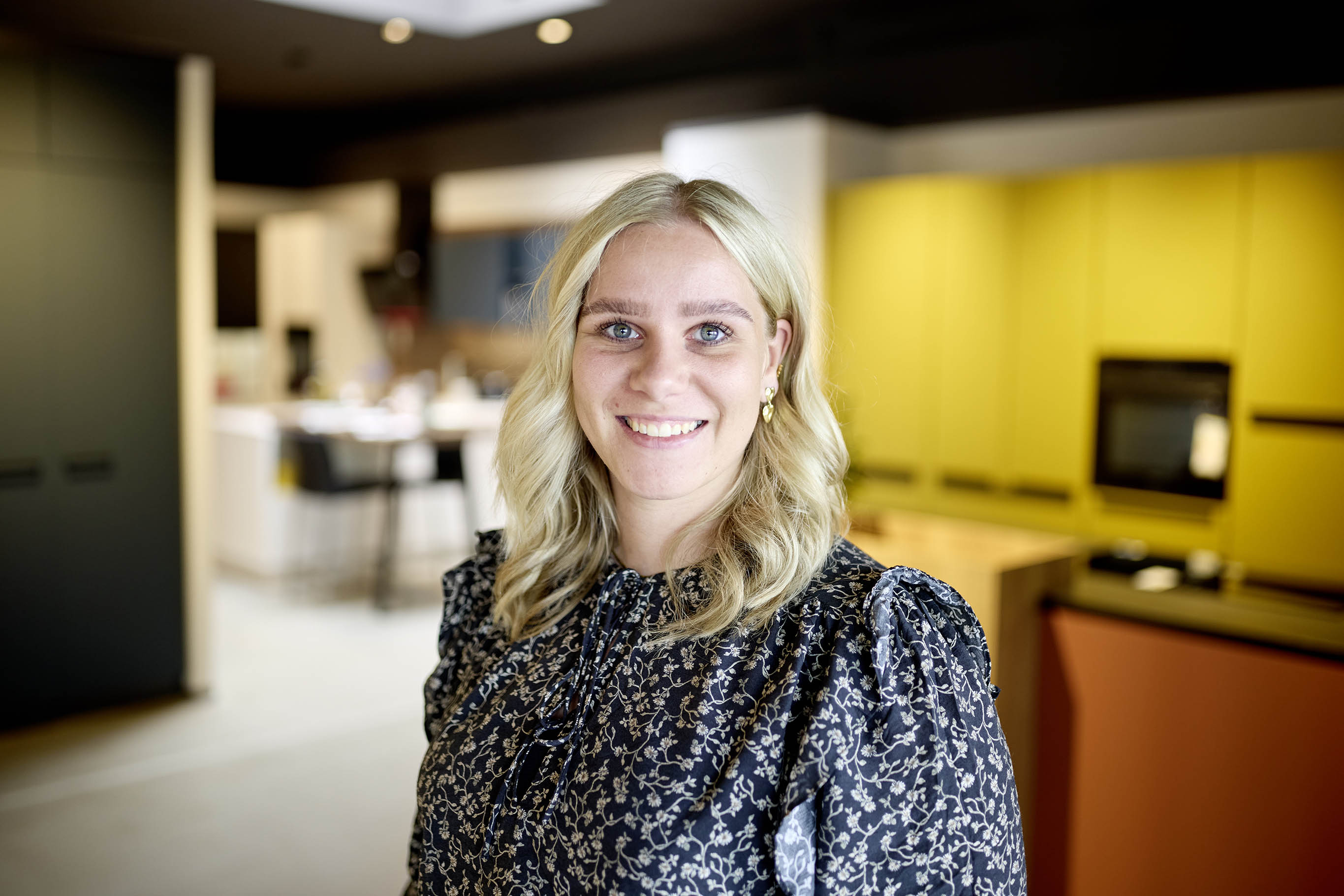 Blonde Frau lächelt in moderner Küche mit gelben Schränken und Holzelementen.
