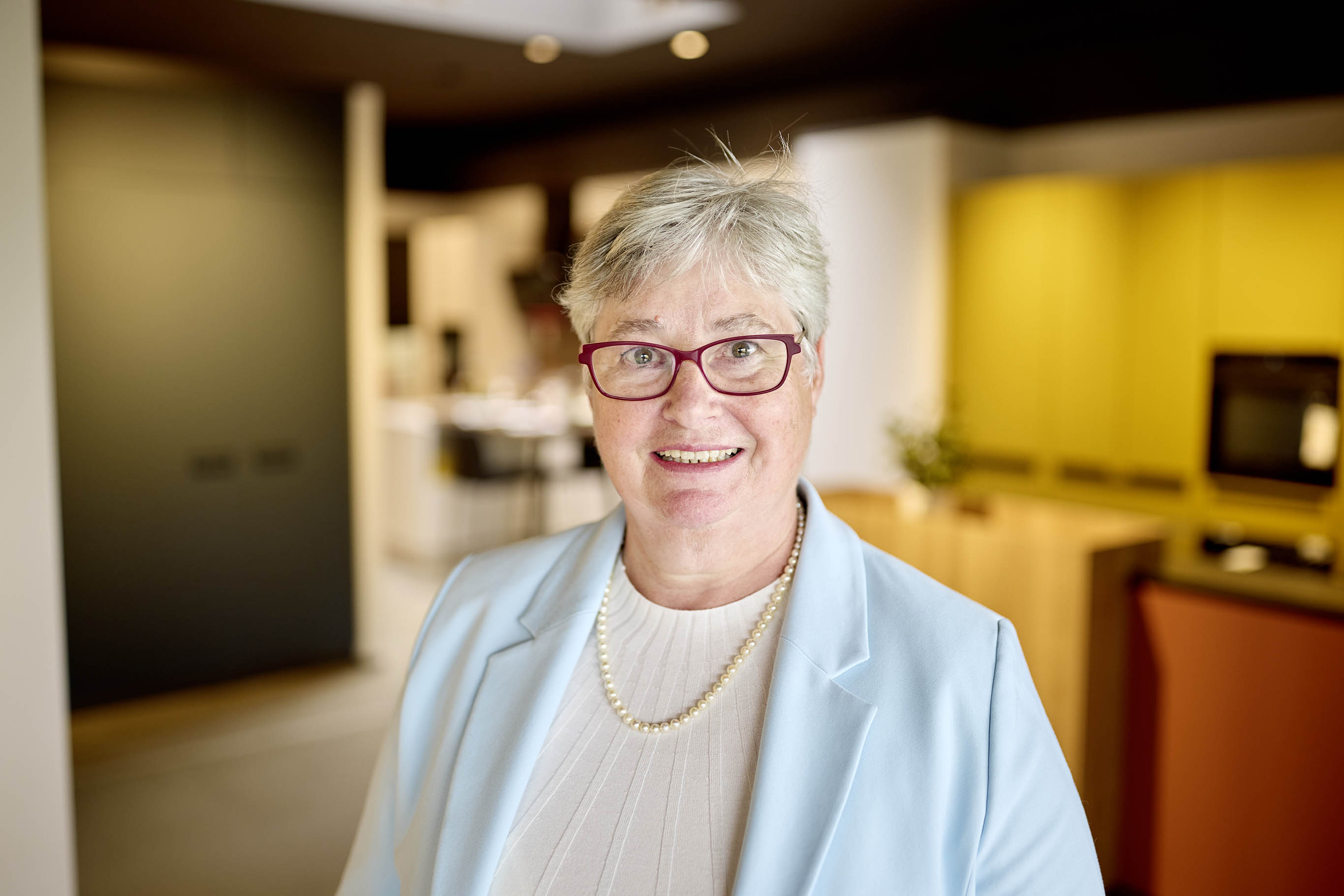 Ältere Dame mit Brille lächelt freundlich, trägt hellblauen Blazer.